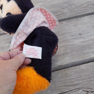 May include: A vintage Minnie Mouse plush toy with a black body, orange feet, and a pink and white polka dot bow. The toy has a tag with text. The plush toy is on a wooden surface.