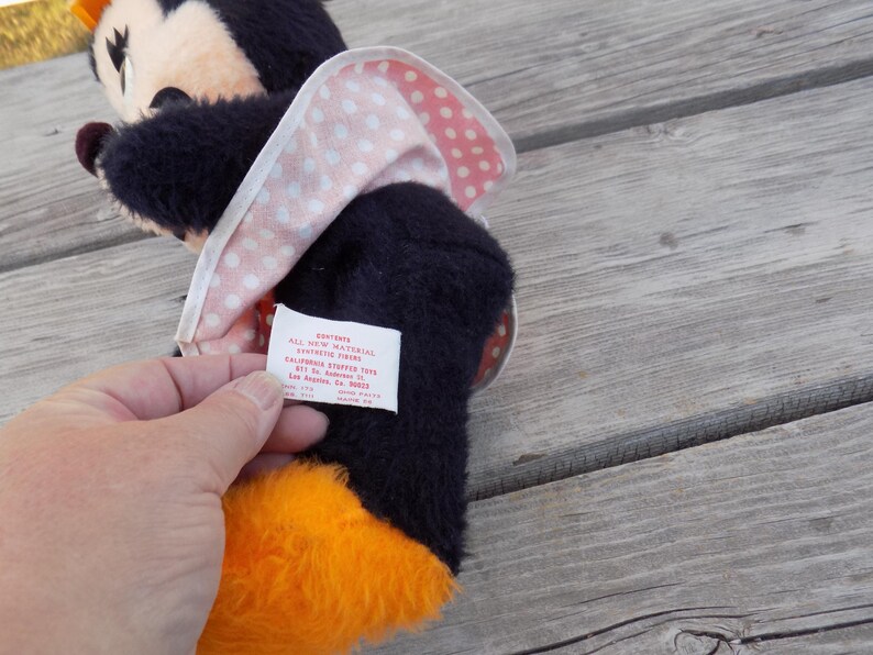 May include: A vintage Minnie Mouse plush toy with a black body, orange feet, and a pink and white polka dot bow. The toy has a tag with text. The plush toy is on a wooden surface.