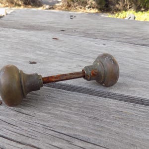 May include: A vintage brass door knob set featuring two spherical knobs connected by a central square metal spindle. The brass surfaces show signs of age and oxidation, with a visible screw on the neck of one knob and a rusted, textured spindle.