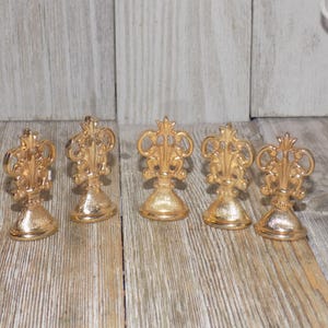 May include: Five gold-coloured metal place card holders with ornate designs. Each holder has a round base and a decorative top.