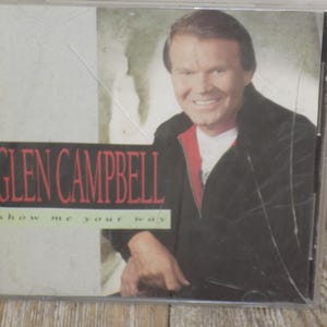 May include: A CD case featuring Glen Campbell. The album cover has a photo of Campbell smiling, wearing a black jacket over a red and white shirt. The album title "Glen Campbell" is in red, and the album subtitle "show me your way" is in white.