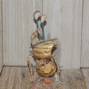 May include: A handcrafted seashell sailboat sculpture. The sculpture features a layered design with beige shells, blue sticks, and small shell accents. The base is a large scallop shell, and the overall aesthetic is nautical.