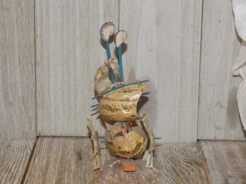 May include: A handcrafted seashell sailboat sculpture. The sculpture features a layered design with beige shells, blue sticks, and small shell accents. The base is a large scallop shell, and the overall aesthetic is nautical.