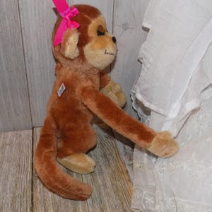 May include: A plush brown monkey with a pink bow on its head. The stuffed animal has tan accents on its face, hands, and feet. It is positioned against a wooden background and white fabric.