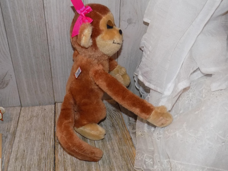 May include: A plush brown monkey with a pink bow on its head. The stuffed animal has tan accents on its face, hands, and feet. It is positioned against a wooden background and white fabric.