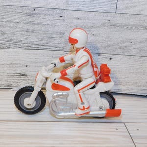 May include: A white and orange toy motorcycle with a matching rider in a helmet and racing suit. The toy has black tires and orange accents. The rider is in a seated position, holding the handlebars.