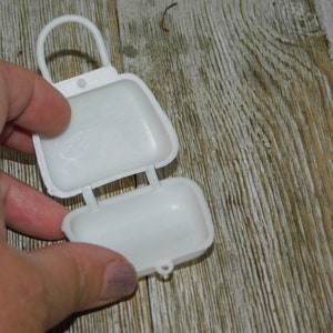 Op de afbeelding: Een witte, scharnierende plastic container, die lijkt op een kleine tas, is open. De container heeft een handvat en wordt tegen een houten achtergrond gehouden.
