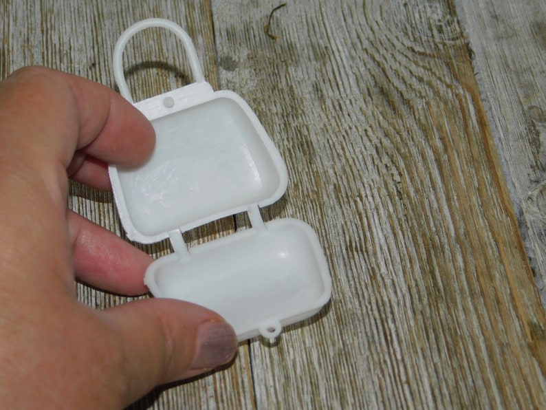 Op de afbeelding: Een witte, scharnierende plastic container, die lijkt op een kleine tas, is open. De container heeft een handvat en wordt tegen een houten achtergrond gehouden.