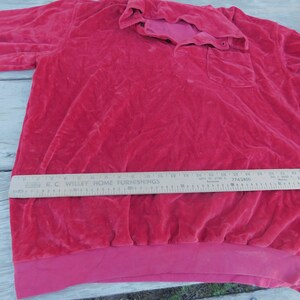 May include: A red velour long-sleeved shirt with a button-up collar and a single pocket on the left side. The shirt has a ribbed hem and is made of a soft, plush fabric.