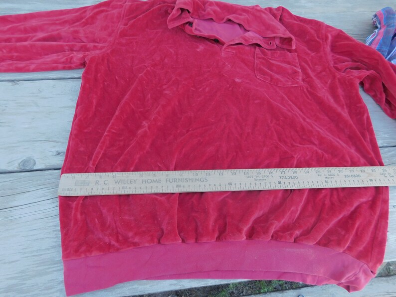 May include: A red velour long-sleeved shirt with a button-up collar and a single pocket on the left side. The shirt has a ribbed hem and is made of a soft, plush fabric.
