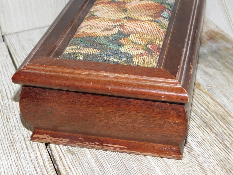 May include: A wooden box with a floral tapestry lid. The box is dark brown and has a slightly rounded shape. The lid is slightly raised and has a decorative border.