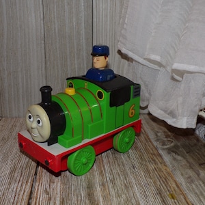 May include: Green toy train with a black face and yellow eyes. The train has a red caboose and a number 6 on the side. A blue conductor figure is sitting in the cab of the train.