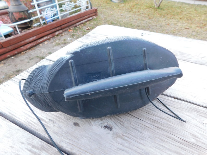 May include: Black plastic decoy of a duck with a black cord attached to the bottom. The decoy has a textured surface and a small black handle on the back.