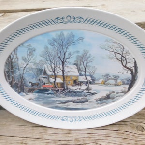 May include: Oval serving platter with a winter scene. The white platter has a blue decorative border. The center features a winter landscape with a cabin, bridge, and horse-drawn carriage. The scene is set in a snowy environment.