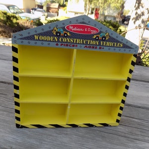 May include: A yellow wooden construction vehicle display shelf with black and yellow stripes. The shelf has six compartments and says "Melissa & Doug Wooden Construction Vehicles 6 Pieces Ages 3+" on the front.