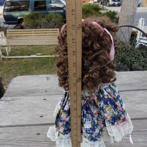 May include: A vintage doll with brown curly hair and a pink ribbon. The doll is wearing a blue floral dress with white lace trim. The doll measures approximately 15 inches in height.