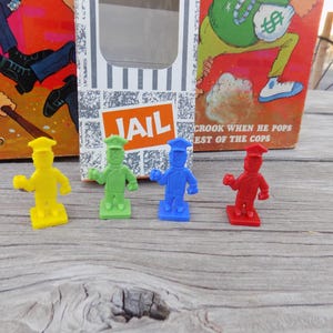 May include: Four colorful plastic game pieces in yellow, green, blue, and red, each depicting a figure in a uniform and hat. The word "JAIL" is visible on a box in the background, along with game artwork.