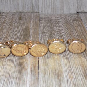 May include: Six gold-coloured wax seals with different designs. The seals are arranged in a row on a wooden surface.