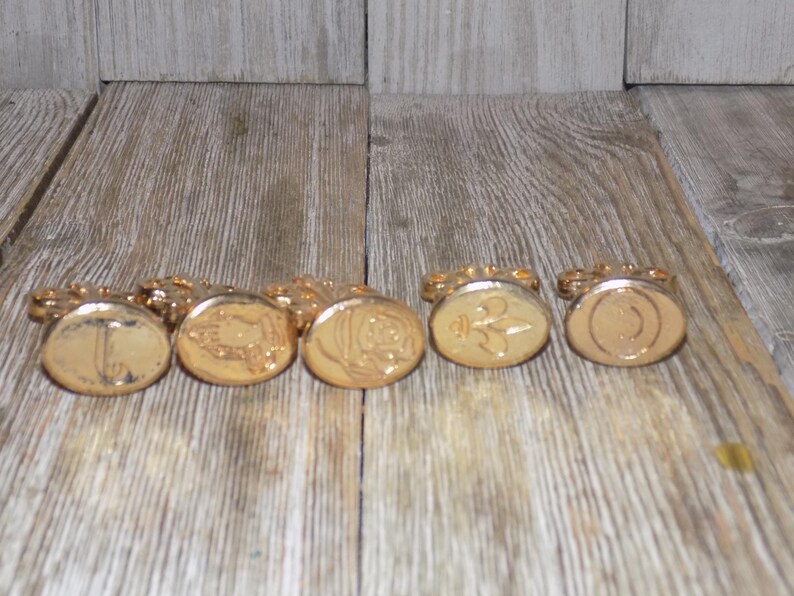 May include: Six gold-coloured wax seals with different designs. The seals are arranged in a row on a wooden surface.