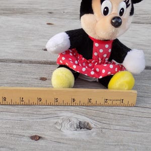 May include: A Minnie Mouse plush toy, featuring a black body, white gloves, and yellow shoes. She wears a red dress with white polka dots. A wooden ruler is in the foreground.