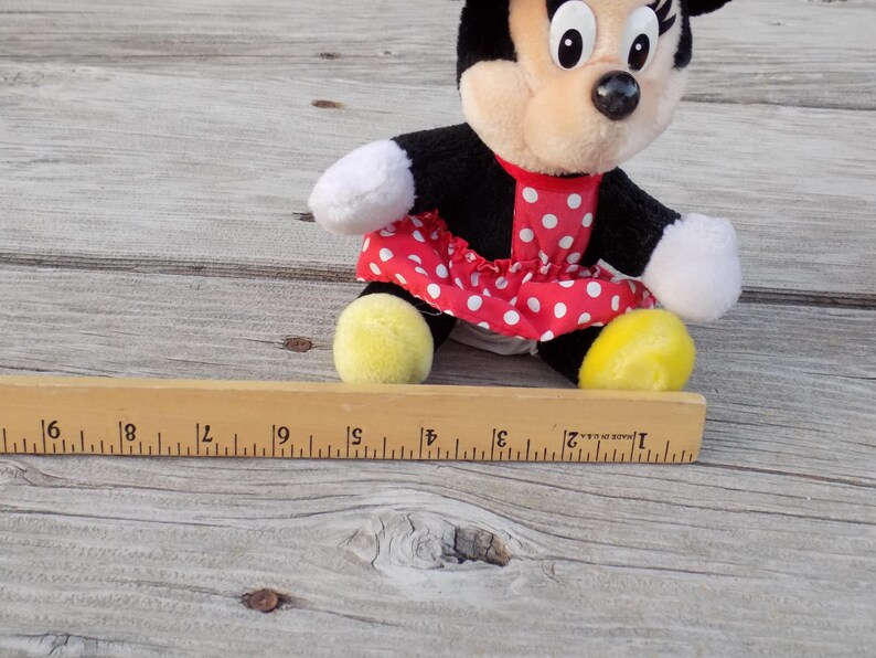 May include: A Minnie Mouse plush toy, featuring a black body, white gloves, and yellow shoes. She wears a red dress with white polka dots. A wooden ruler is in the foreground.
