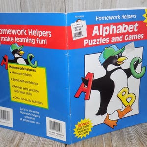 May include: A blue and red Homework Helpers book titled "Alphabet Puzzles and Games" for grades K-1. The cover features a cartoon penguin with the letters A, B, and C. The book promotes learning with fun activities.