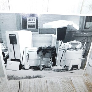 May include: A black and white photo of a row of appliances outside a building. The appliances include a white refrigerator, a white washing machine, and a white dryer. A shopping cart is in front of the washing machine. A sign that reads 
