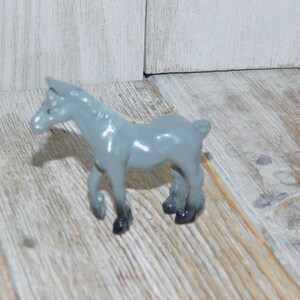 May include: A gray plastic toy horse figurine with black hooves. The horse is standing on a light wood surface.