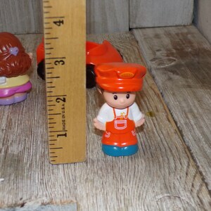 May include: A set of three plastic figurines, including a girl with brown hair wearing a yellow shirt and pink trousers, a boy wearing a red hat and overalls, and a red car. The figurines are approximately 5 cm tall.