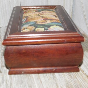 May include: A wooden box with a hinged lid. The lid is covered in a tapestry fabric with a floral pattern. The box is dark brown and has a slightly rounded shape.
