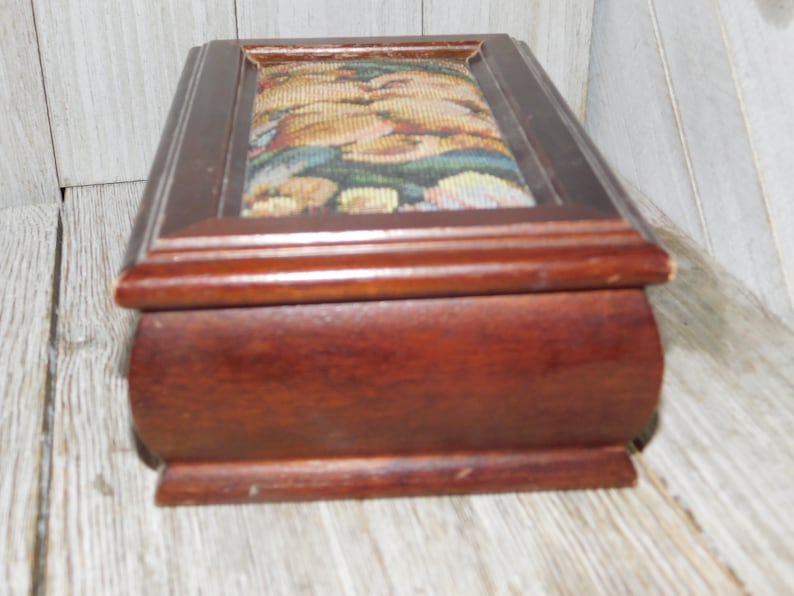 May include: A wooden box with a hinged lid. The lid is covered in a tapestry fabric with a floral pattern. The box is dark brown and has a slightly rounded shape.