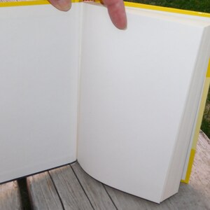 May include: A yellow hardback book with white pages is open to a blank page. The book is lying on a wooden surface.