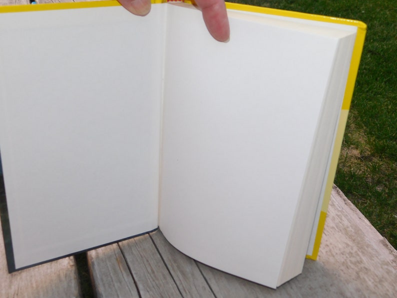 May include: A yellow hardback book with white pages is open to a blank page. The book is lying on a wooden surface.