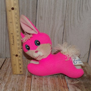 May include: A pink plush bunny rabbit toy with white fur accents and blue eyes. The toy is lying on a wooden surface and has a white tag that says "Casti" on it.