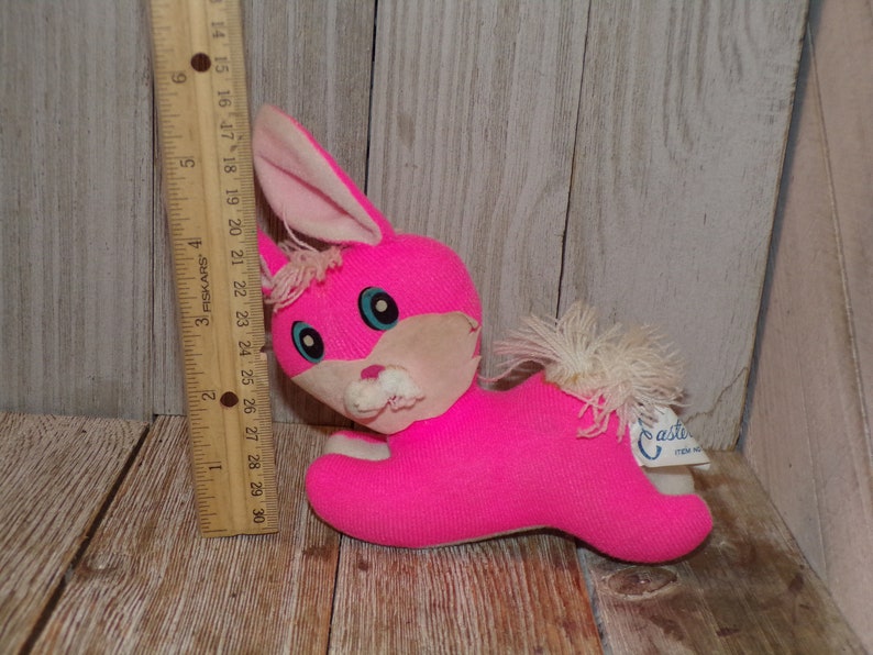 May include: A pink plush bunny rabbit toy with white fur accents and blue eyes. The toy is lying on a wooden surface and has a white tag that says "Casti" on it.