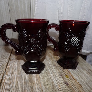 May include: Two ruby red glass mugs with ornate handles and detailed floral patterns. The mugs have a pedestal base and are displayed on a wooden surface. The mugs are approximately 4 inches tall.