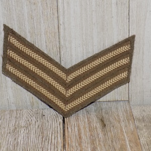 May include: A brown chevron-shaped patch with three parallel, light-colored, chevron-patterned stripes. The patch is made of a textured fabric and is set against a wooden background.