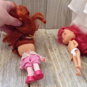 Pode incluir: Duas bonecas de cabelo ruivo, uma com um vestido rosa e a outra com roupa interior branca. As bonecas t&ecirc;m rostos pintados e est&atilde;o deitadas sobre uma superf&iacute;cie de madeira.