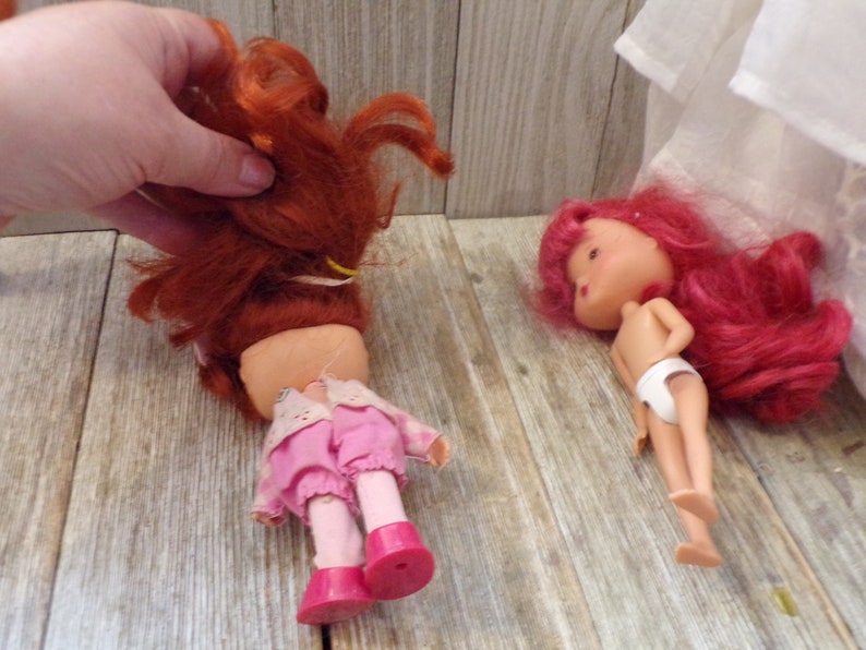 Pode incluir: Duas bonecas de cabelo ruivo, uma com um vestido rosa e a outra com roupa interior branca. As bonecas t&ecirc;m rostos pintados e est&atilde;o deitadas sobre uma superf&iacute;cie de madeira.