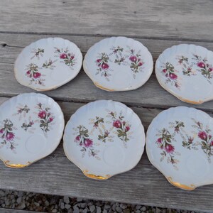 May include: Six white porcelain saucers with a scalloped edge and gold trim. Each saucer features a floral design with pink roses and green leaves. The saucers are arranged on a weathered wooden surface.