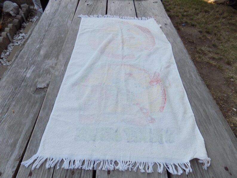 Peut inclure: Serviette rectangulaire blanche avec un motif color&eacute;. La serviette a un bord &agrave; franges et semble &ecirc;tre faite d'un mat&eacute;riau doux et absorbant. Le motif pr&eacute;sente des &eacute;l&eacute;ments rouges, jaunes et bleus. La serviette est pos&eacute;e sur une surface en bois vieilli.