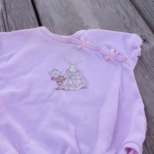 May include: A pink baby onesie with a white rabbit illustration wearing a dress and holding a teacup. The onesie has pink bows and buttons on the shoulder.