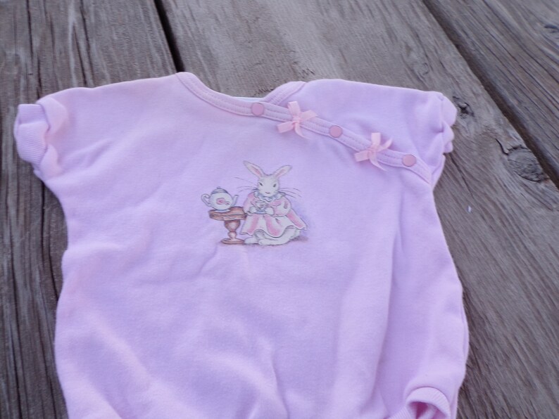 May include: A pink baby onesie with a white rabbit illustration wearing a dress and holding a teacup. The onesie has pink bows and buttons on the shoulder.