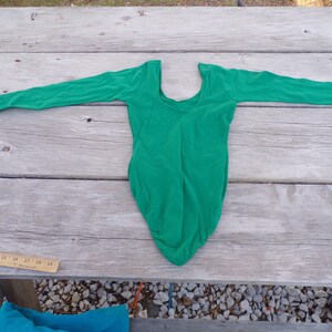 Puede incluir: Un leotardo verde de manga larga con cuello redondo. El leotardo est&aacute; colocado plano sobre una superficie de madera.