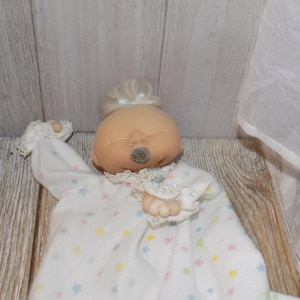 May include: A soft, plush baby doll with a light skin tone, closed eyes, and a small, gray nose. The doll wears a white onesie patterned with pastel stars and trimmed with lace. A white bow adorns the doll's light-colored hair.
