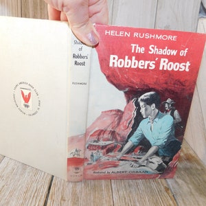 May include: The cover of a paperback book titled "The Shadow of Robbers' Roost" by Helen Rushmore. The cover features a young boy in a blue shirt looking out from behind a red rock formation. The book is illustrated by Albert Orbaan.