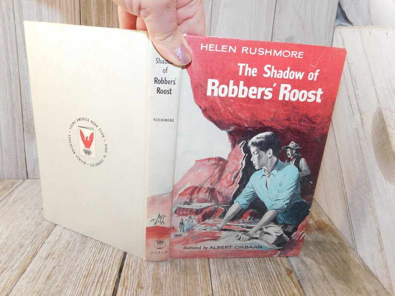 May include: The cover of a paperback book titled "The Shadow of Robbers' Roost" by Helen Rushmore. The cover features a young boy in a blue shirt looking out from behind a red rock formation. The book is illustrated by Albert Orbaan.