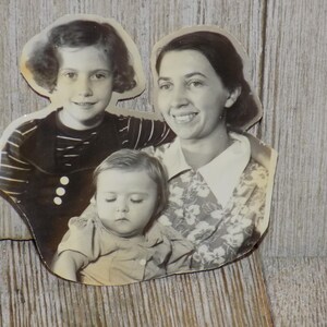 May include: Vintage black and white photograph featuring three people. A young girl in a striped top and a woman with a floral print blouse are smiling. A baby is nestled in the woman's arms. The photo is cut around the figures.