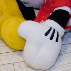 Op de afbeelding: Close-up van een pluche speelgoed met een witte handschoen met zwarte details, een gele voet en een rode mouw met stippen. Het speelgoed is waarschijnlijk een personage uit een populaire tekenfilm.