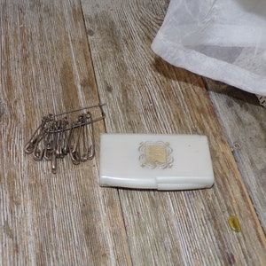 May include: A white plastic case with a gold design and a pile of safety pins on a wooden surface.